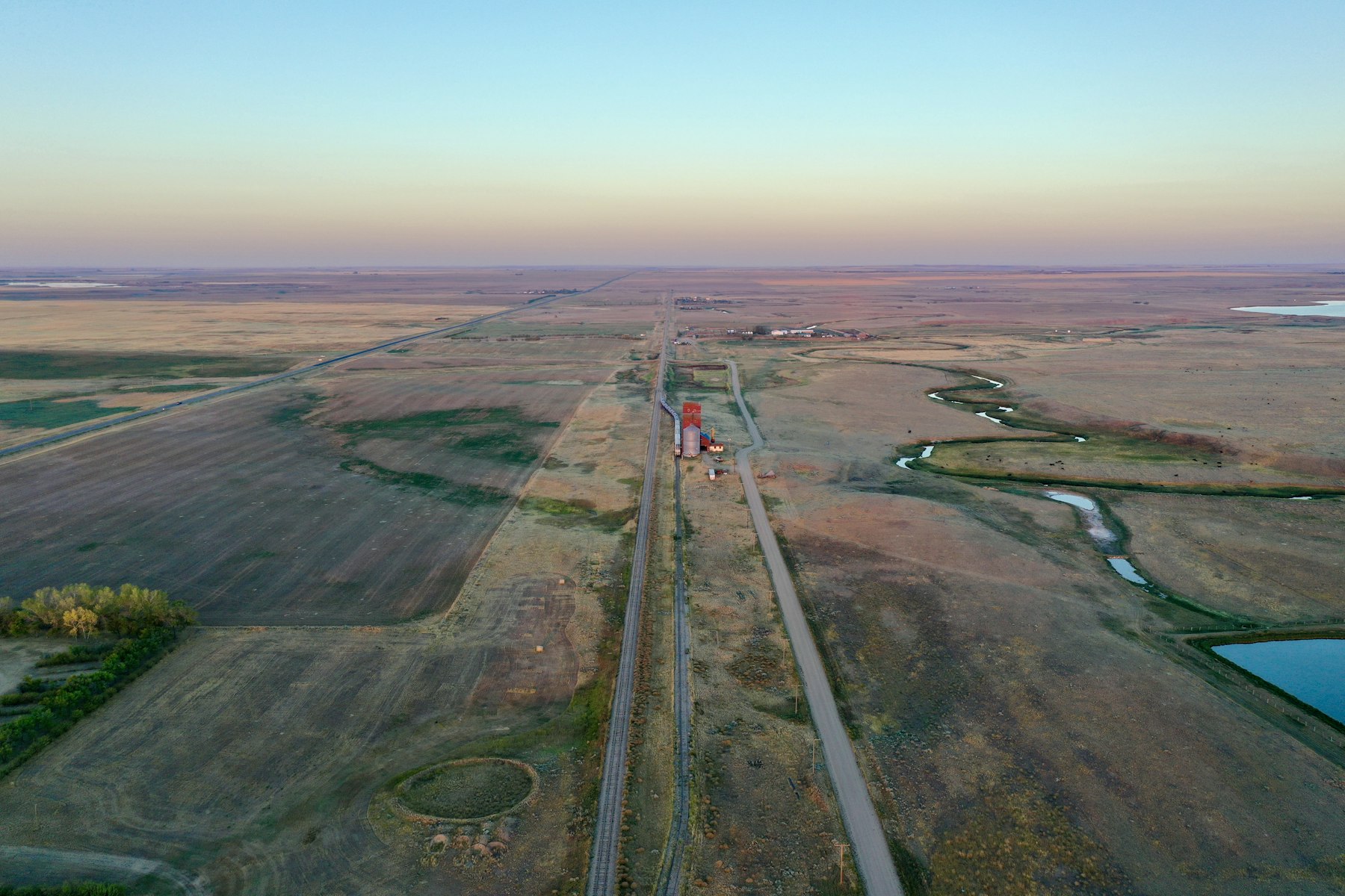 Prairie farmland at dusk with a rail line, a road and a grain elevator